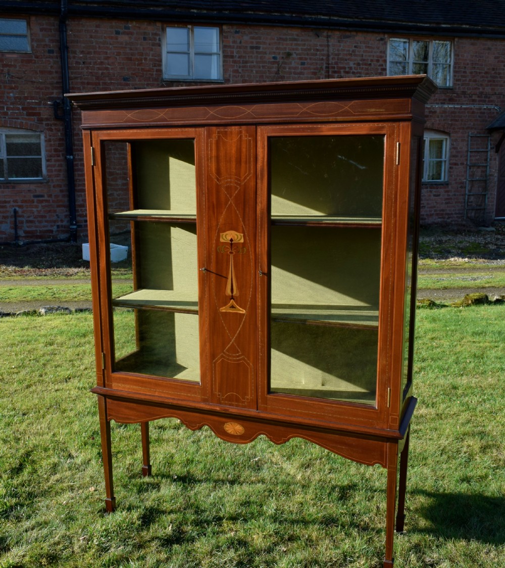 An early 20th-century mahogany Art Nouveau display cabinet featuring glazed doors, central inlaid botanical marquetry, and a fabric-lined interior on slender legs. View VI