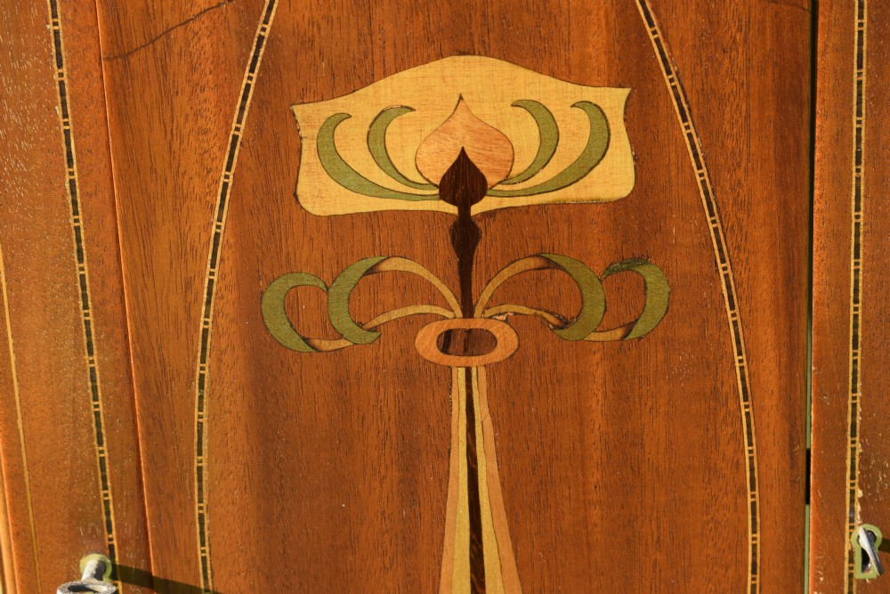 An early 20th-century mahogany Art Nouveau display cabinet featuring glazed doors, central inlaid botanical marquetry, and a fabric-lined interior on slender legs. View XV