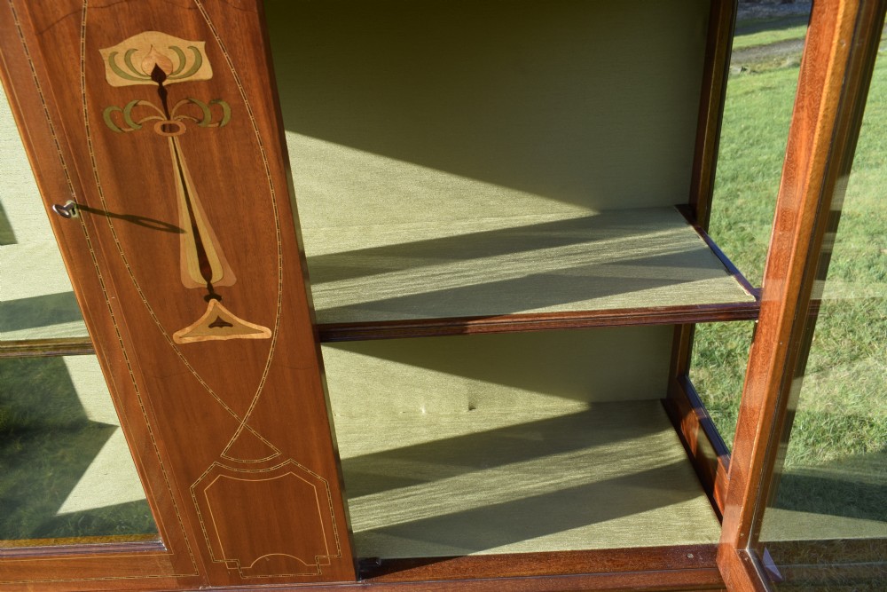 An early 20th-century mahogany Art Nouveau display cabinet featuring glazed doors, central inlaid botanical marquetry, and a fabric-lined interior on slender legs. View XXI