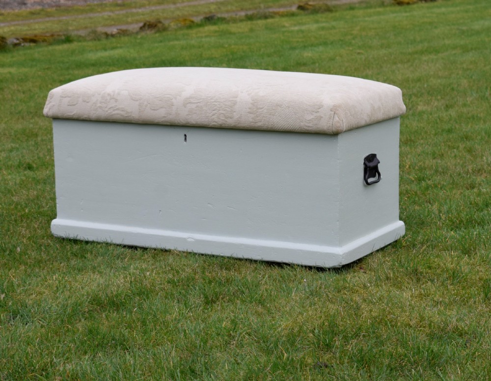 A Victorian painted pine blanket box featuring a cream upholstered cushioned top and iron drop side handles.