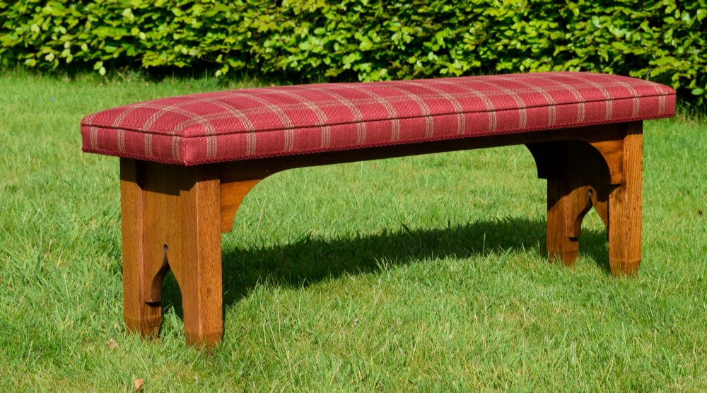 An oak window bench featuring a padded red tartan upholstered seat supported by solid oak legs with Gothic arch cut-out details. View II