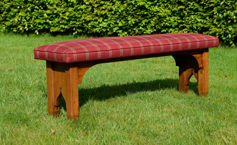 An oak window bench featuring a padded red tartan upholstered seat supported by solid oak legs with Gothic arch cut-out details. View III