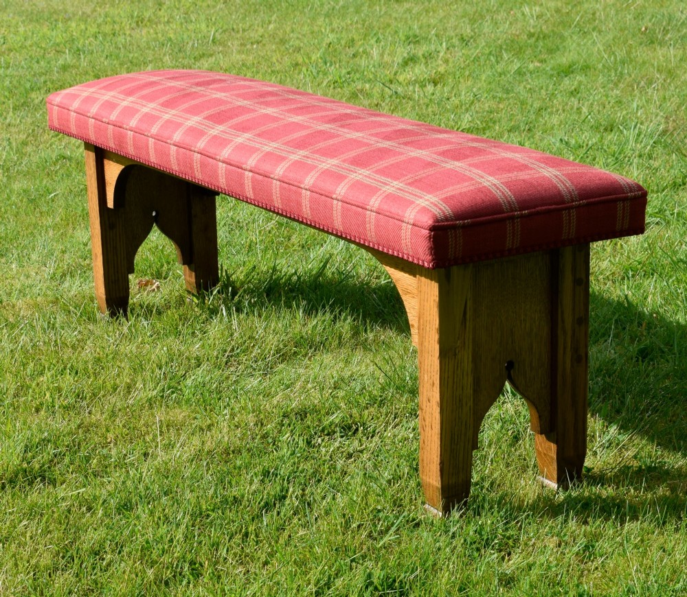 An oak window bench featuring a padded red tartan upholstered seat supported by solid oak legs with Gothic arch cut-out details. View V
