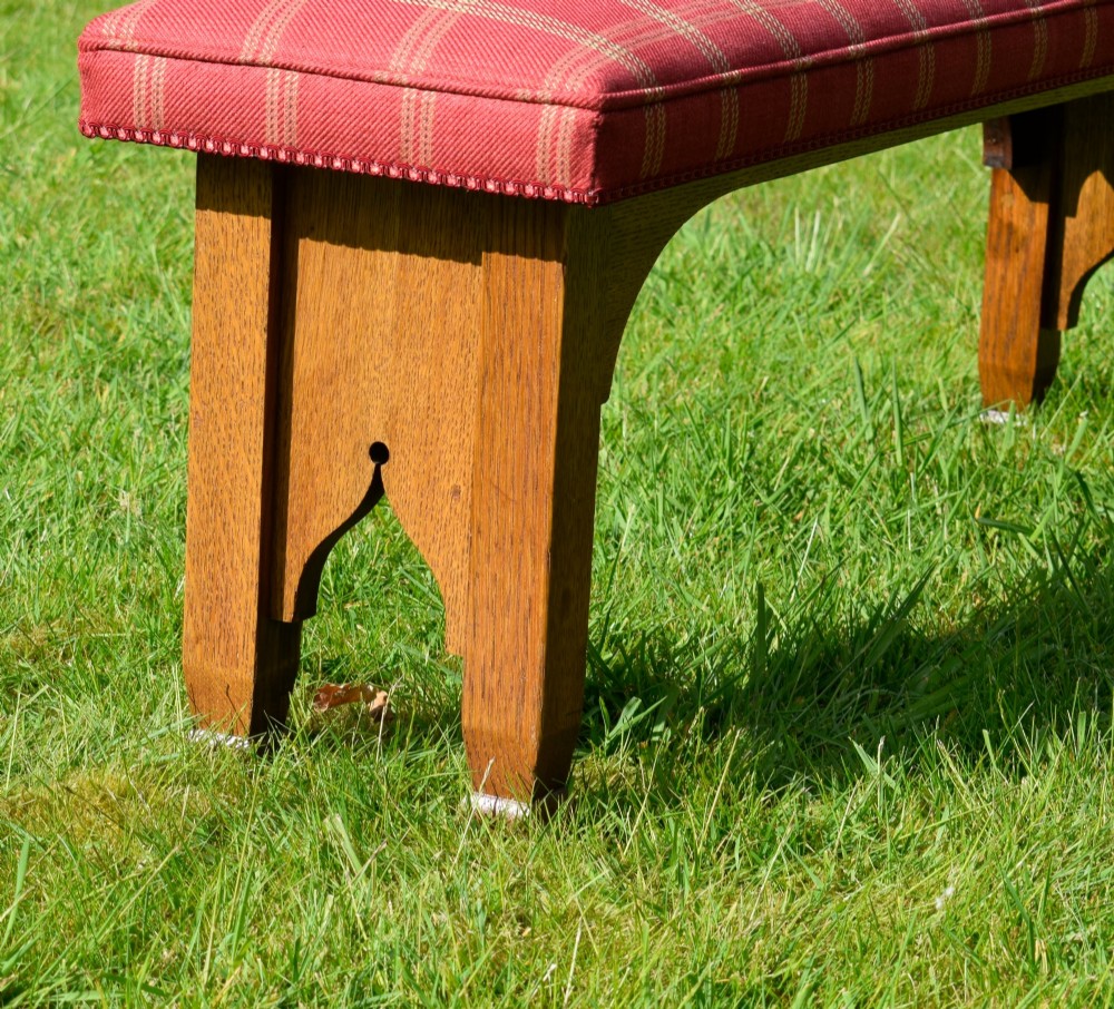 An oak window bench featuring a padded red tartan upholstered seat supported by solid oak legs with Gothic arch cut-out details. View VI