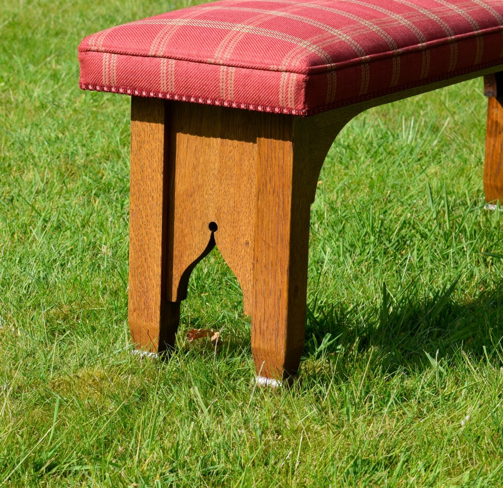 An oak window bench featuring a padded red tartan upholstered seat supported by solid oak legs with Gothic arch cut-out details. View VII
