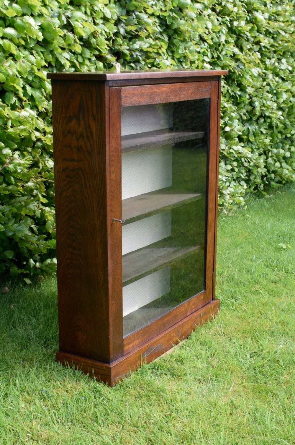 A dark oak glazed cabinet featuring a single glass door opening to internal shelving backed with a neutral patterned fabric lining. View II