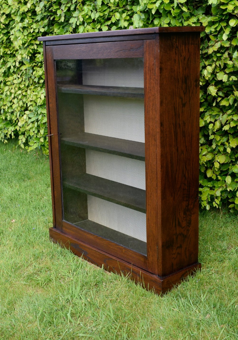 A dark oak glazed cabinet featuring a single glass door opening to internal shelving backed with a neutral patterned fabric lining. View III