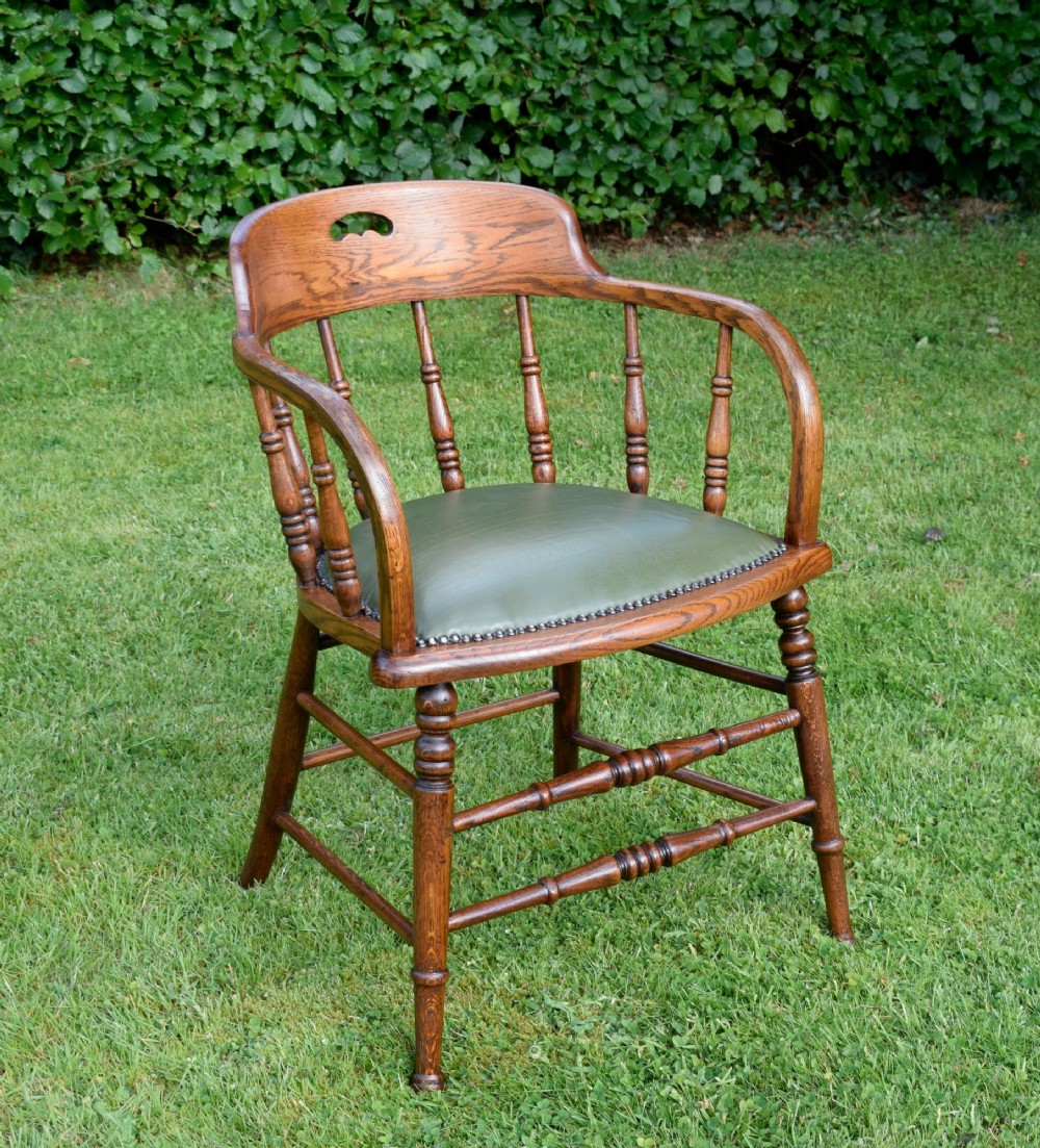 An early 20th-century oak desk chair featuring turned spindles, a curved backrest with a cut-out handle, and a green leather seat fastened with brass studs.