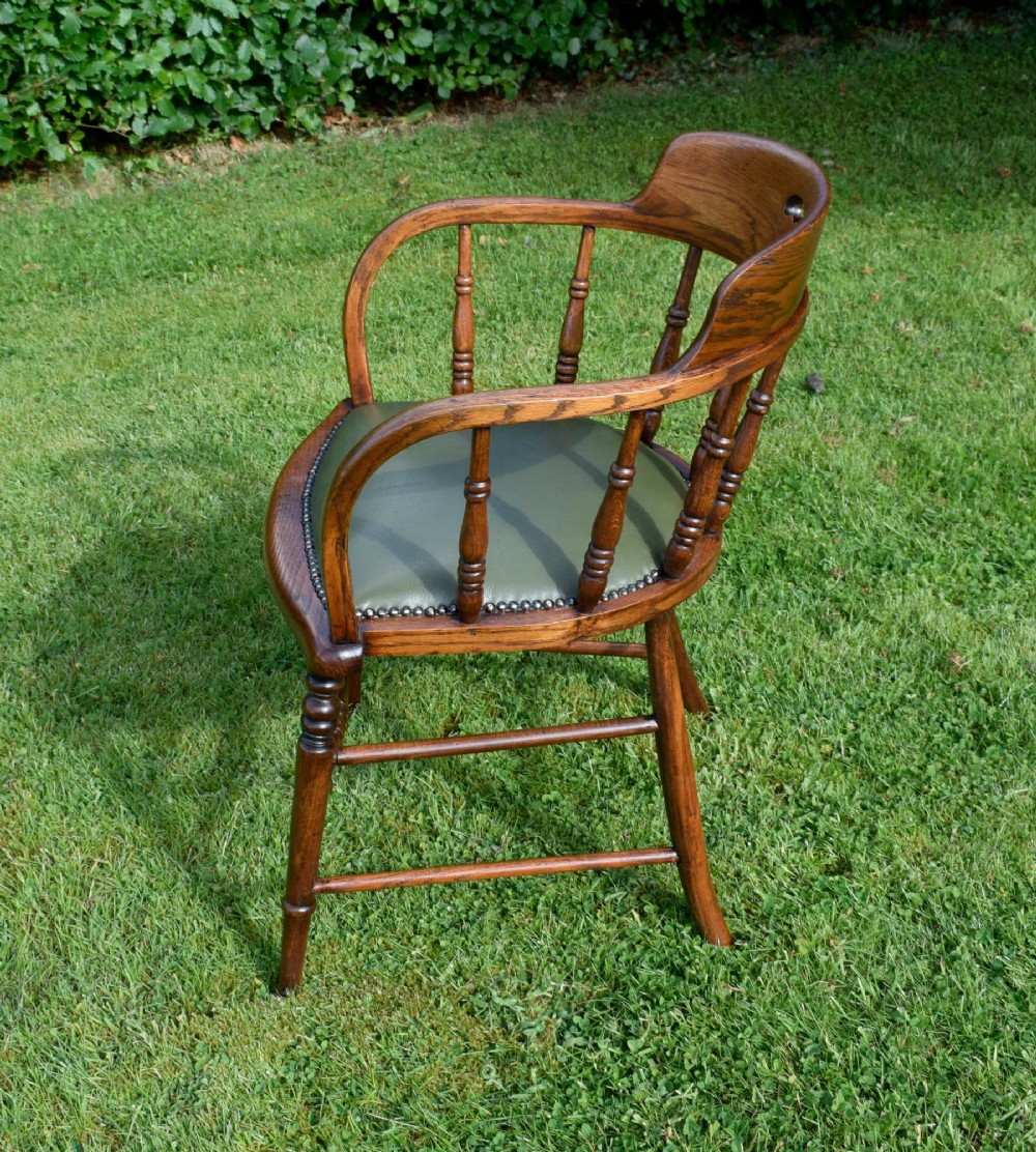 An early 20th-century oak desk chair featuring turned spindles, a curved backrest with a cut-out handle, and a green leather seat fastened with brass studs. View VI