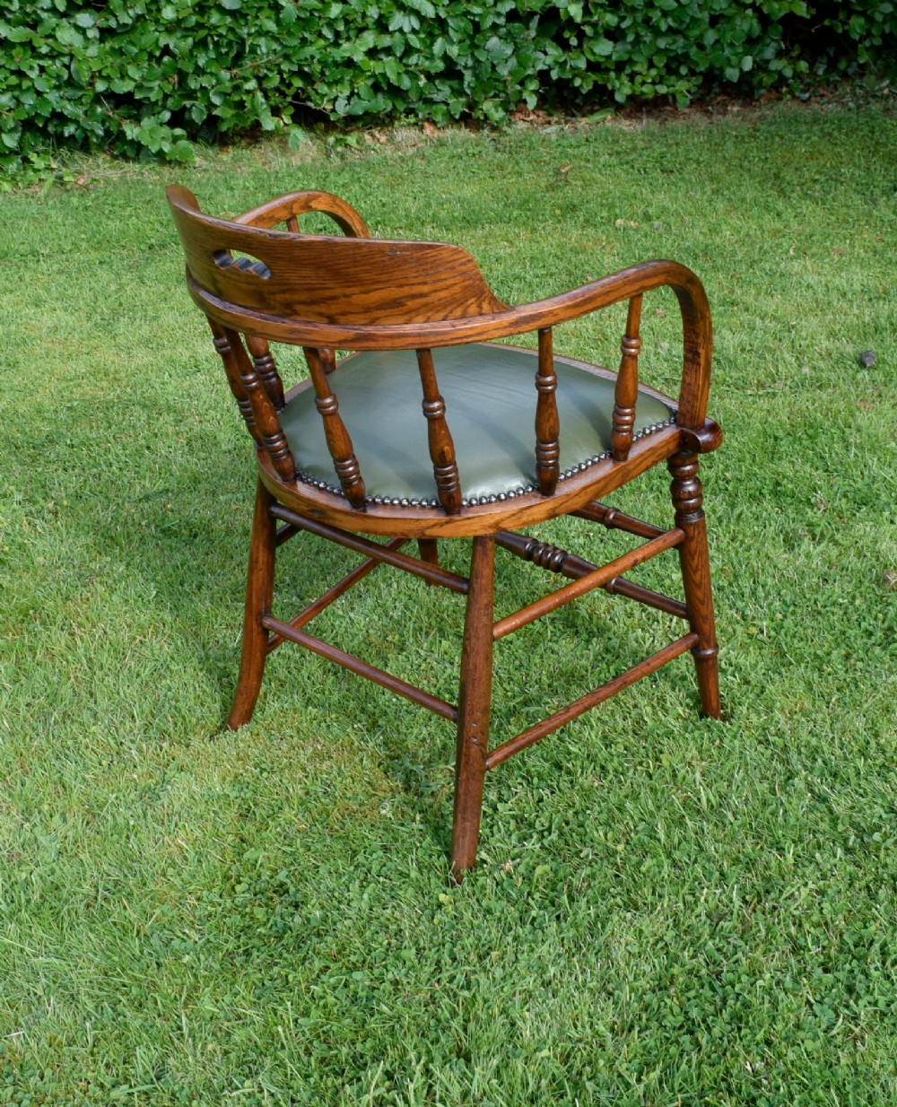 An early 20th-century oak desk chair featuring turned spindles, a curved backrest with a cut-out handle, and a green leather seat fastened with brass studs. View VII