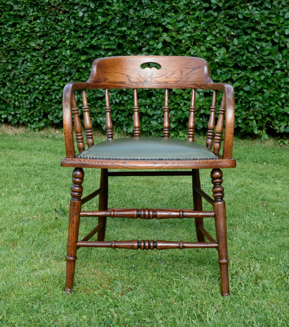 An early 20th-century oak desk chair featuring turned spindles, a curved backrest with a cut-out handle, and a green leather seat fastened with brass studs. View VIII