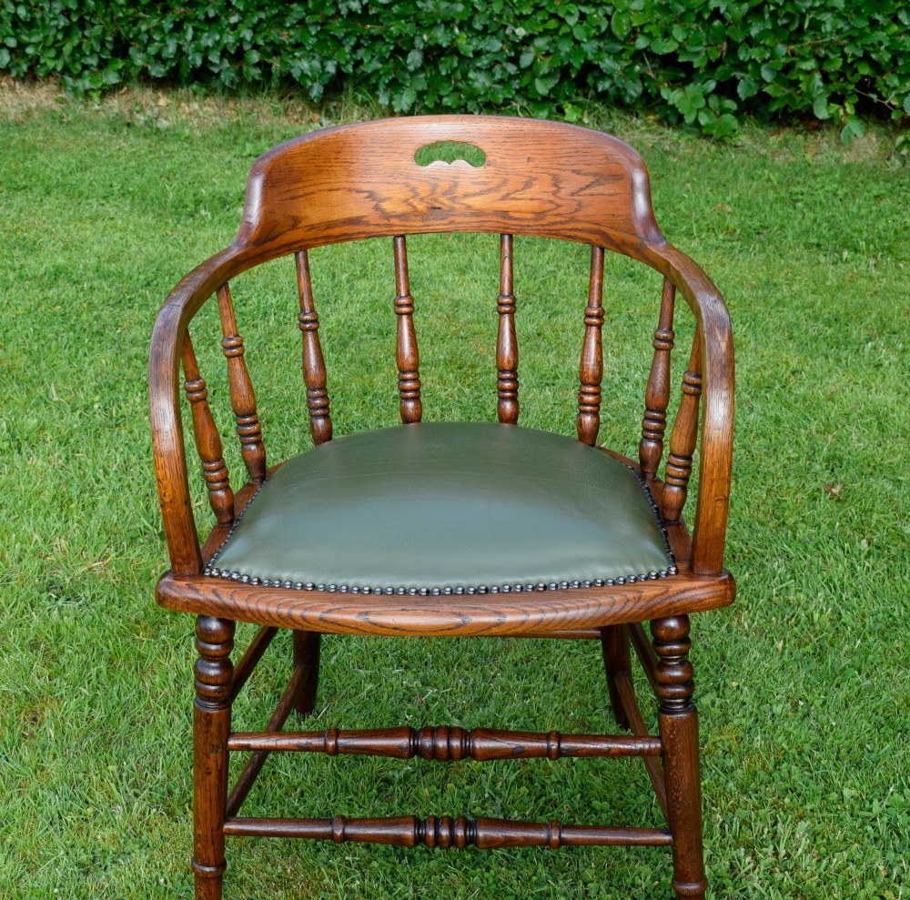 An early 20th-century oak desk chair featuring turned spindles, a curved backrest with a cut-out handle, and a green leather seat fastened with brass studs. View IX