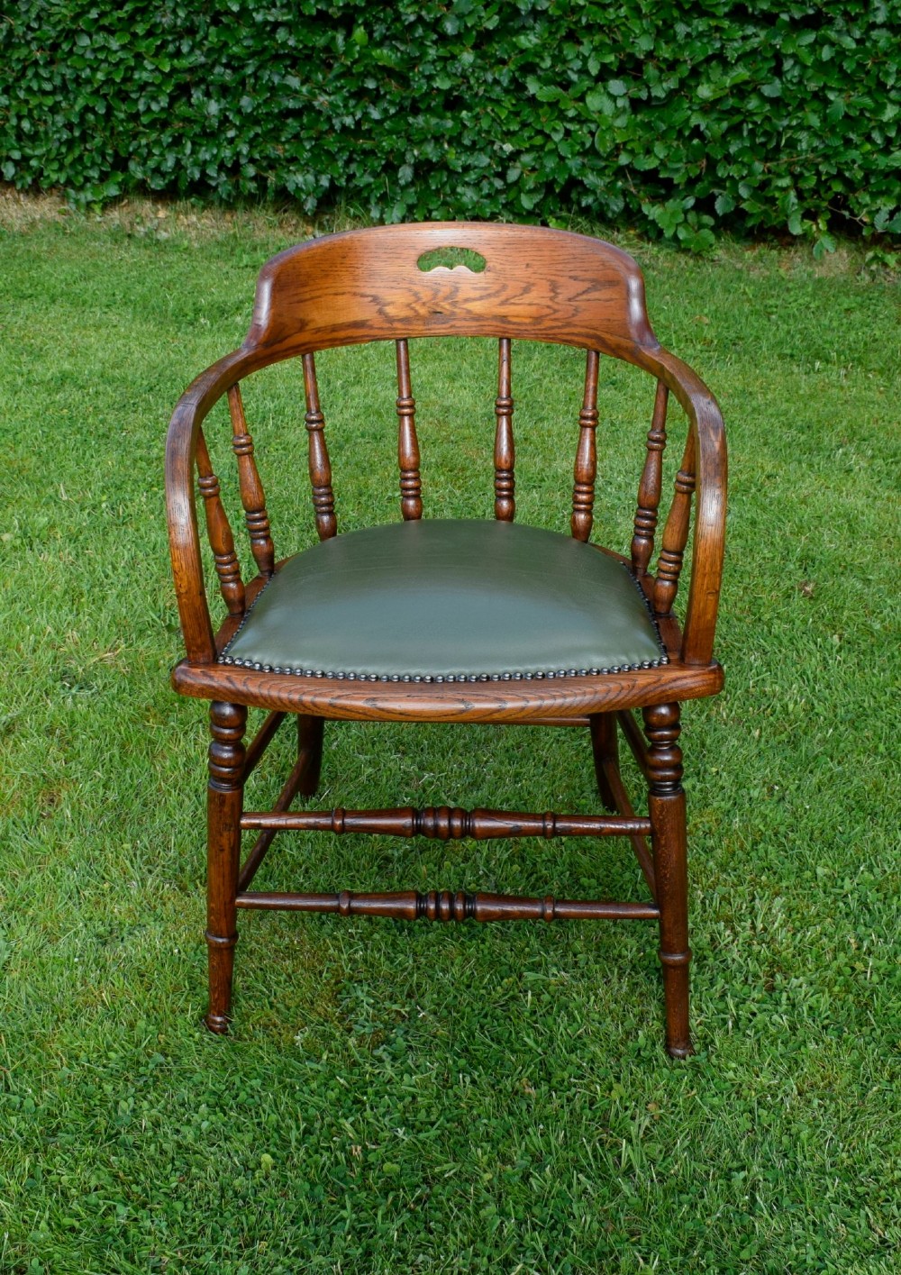 An early 20th-century oak desk chair featuring turned spindles, a curved backrest with a cut-out handle, and a green leather seat fastened with brass studs. View X