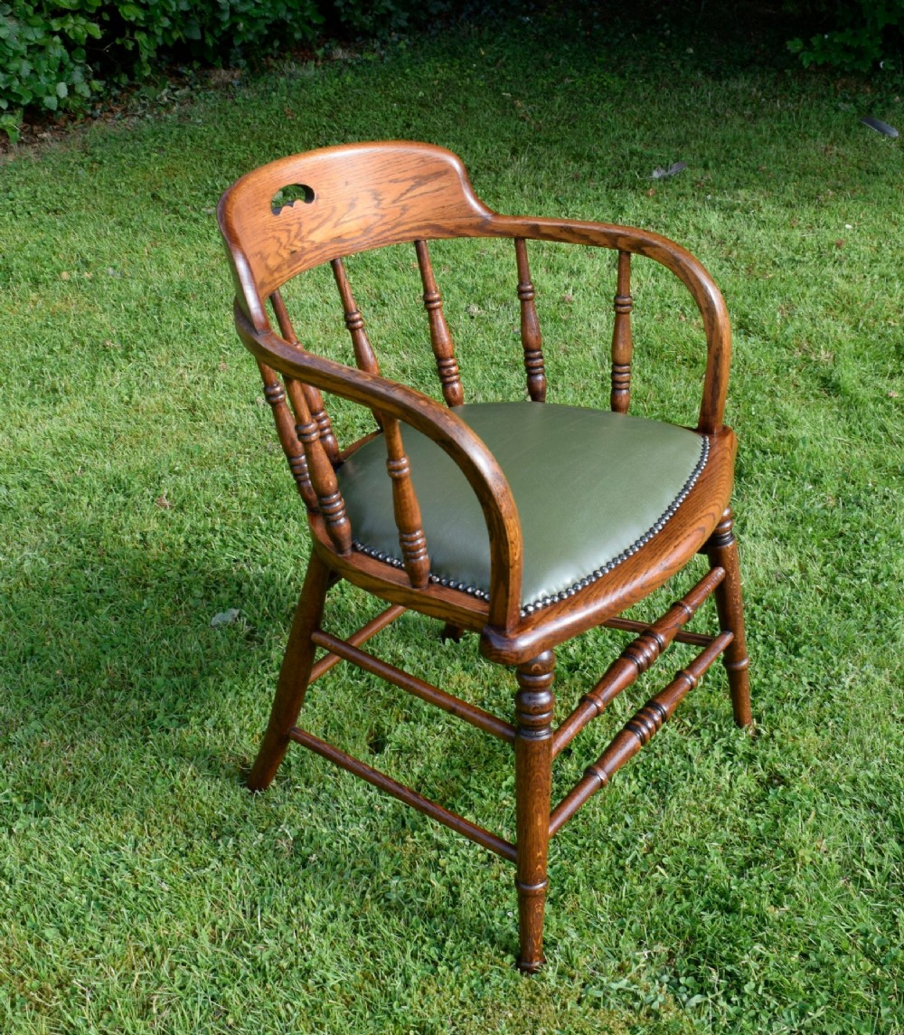 An early 20th-century oak desk chair featuring turned spindles, a curved backrest with a cut-out handle, and a green leather seat fastened with brass studs. View XI