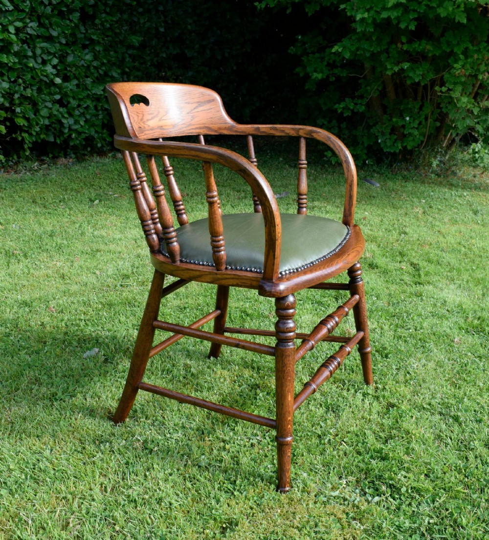 An early 20th-century oak desk chair featuring turned spindles, a curved backrest with a cut-out handle, and a green leather seat fastened with brass studs. View XII