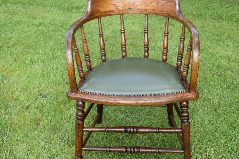An early 20th-century oak desk chair featuring turned spindles, a curved backrest with a cut-out handle, and a green leather seat fastened with brass studs. View XIV