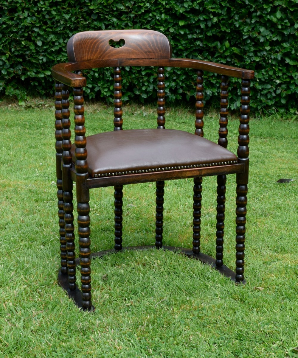 A dark oak Arts and Crafts desk chair featuring bobbin-turned uprights, a curved crest rail with a cut-out handle, and a brown leather seat fastened with brass studs.