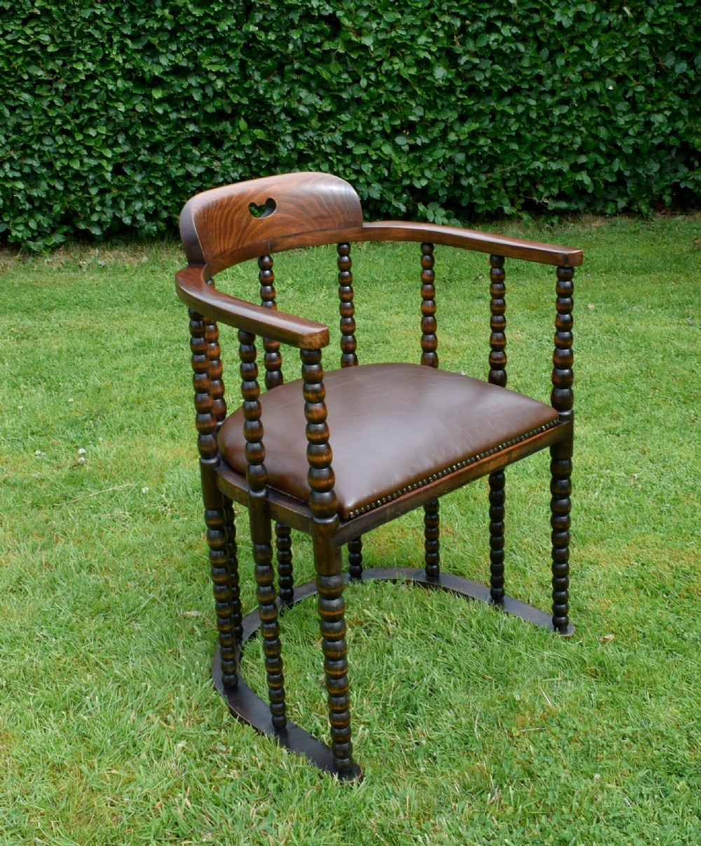 A dark oak Arts and Crafts desk chair featuring bobbin-turned uprights, a curved crest rail with a cut-out handle, and a brown leather seat fastened with brass studs. View V