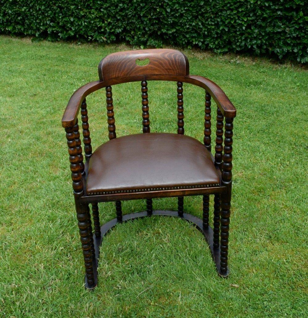 A dark oak Arts and Crafts desk chair featuring bobbin-turned uprights, a curved crest rail with a cut-out handle, and a brown leather seat fastened with brass studs. View VI