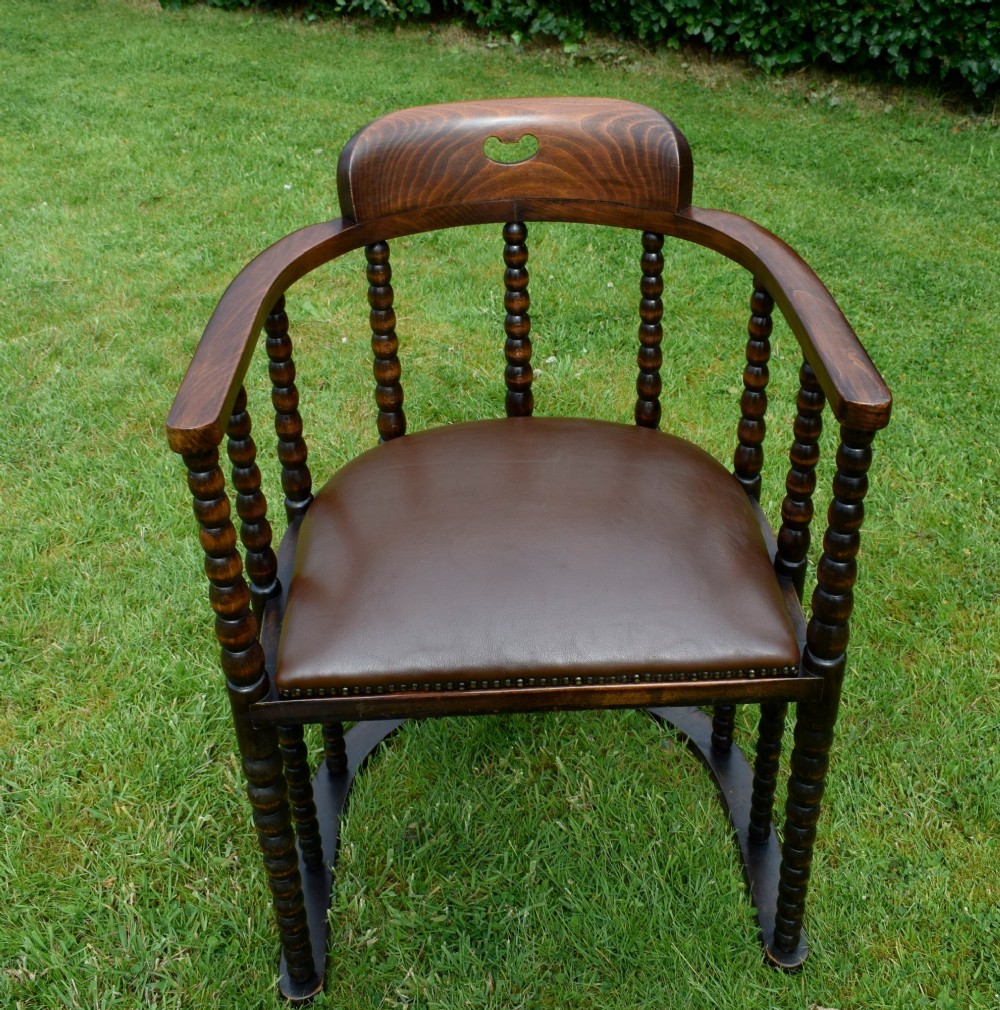 A dark oak Arts and Crafts desk chair featuring bobbin-turned uprights, a curved crest rail with a cut-out handle, and a brown leather seat fastened with brass studs. View VII