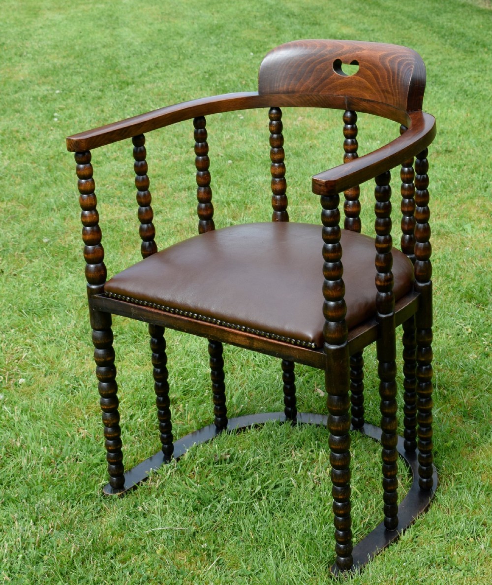 A dark oak Arts and Crafts desk chair featuring bobbin-turned uprights, a curved crest rail with a cut-out handle, and a brown leather seat fastened with brass studs. View X