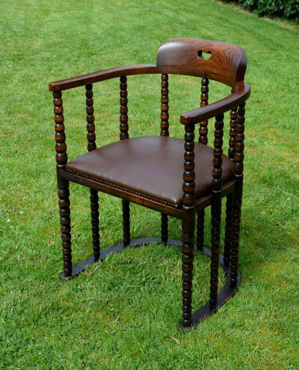 A dark oak Arts and Crafts desk chair featuring bobbin-turned uprights, a curved crest rail with a cut-out handle, and a brown leather seat fastened with brass studs. View XII
