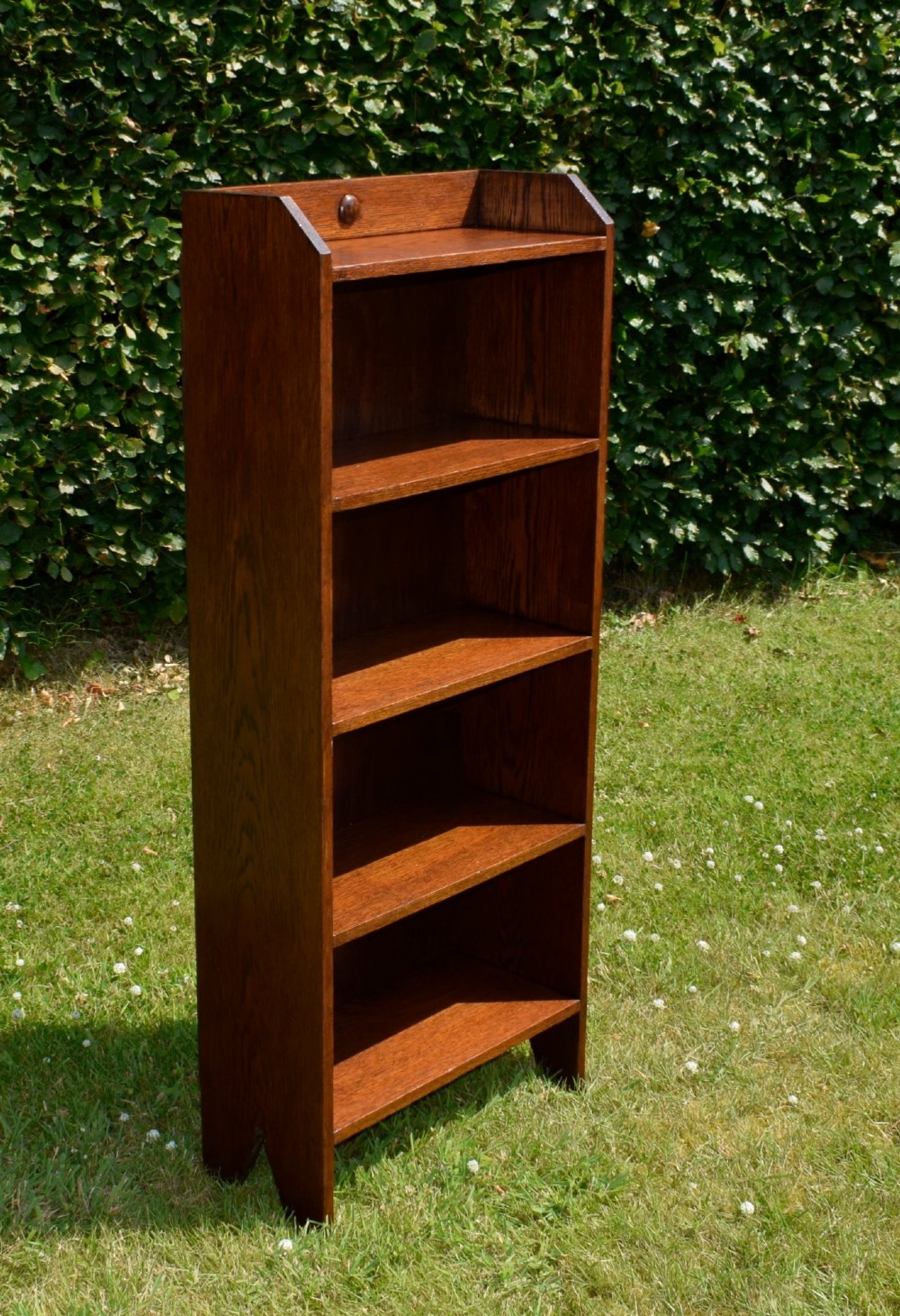 A slim, open-front Arts and Crafts oak bookcase with five shelves, cut-out side panels, and a low galleried top featuring a subtle central carved motif.
