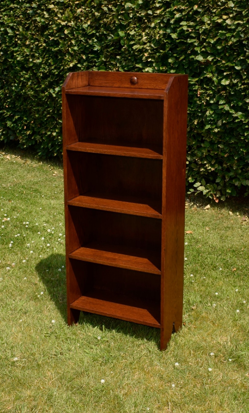A slim, open-front Arts and Crafts oak bookcase with five shelves, cut-out side panels, and a low galleried top featuring a subtle central carved motif. View II