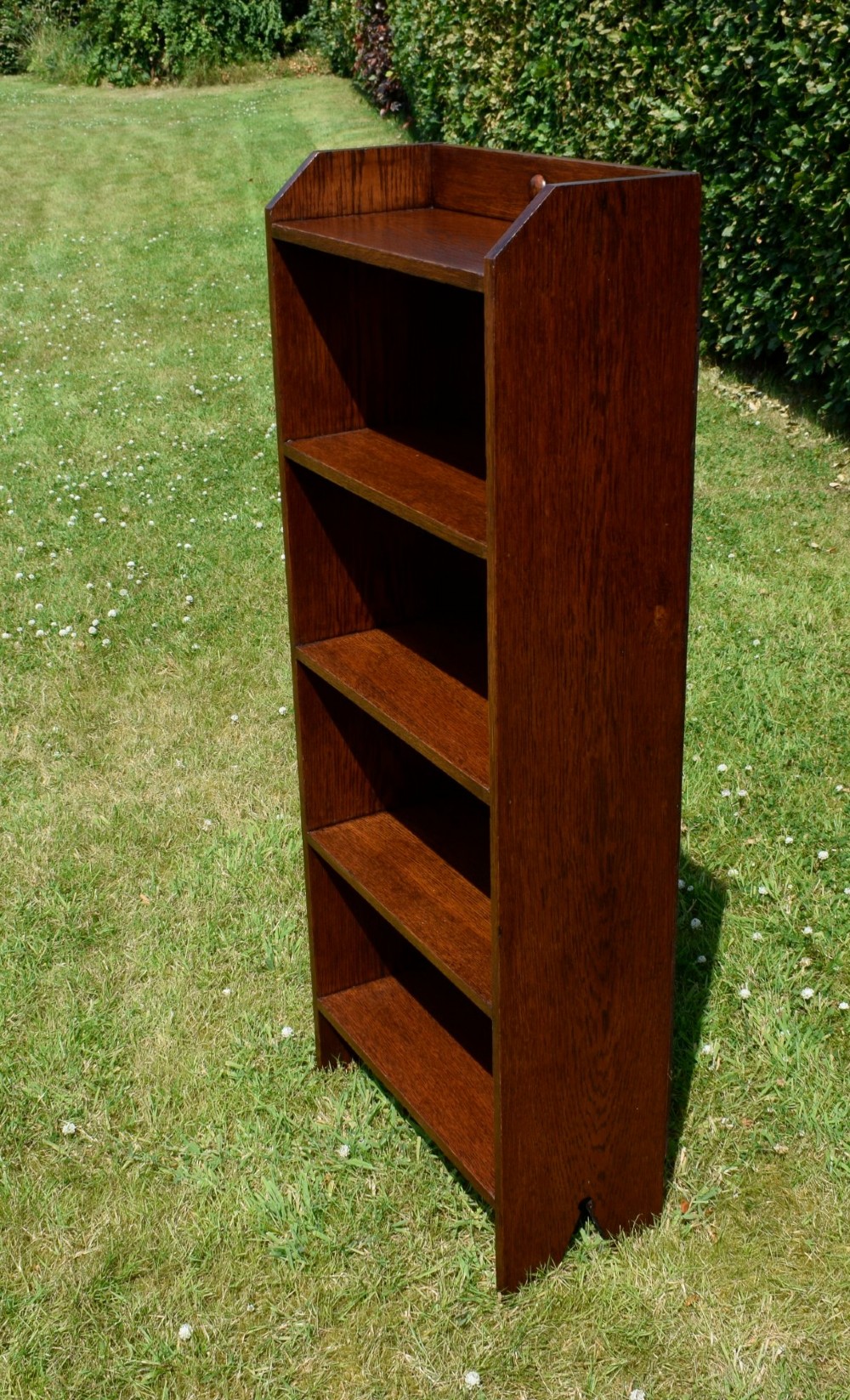 A slim, open-front Arts and Crafts oak bookcase with five shelves, cut-out side panels, and a low galleried top featuring a subtle central carved motif. View III
