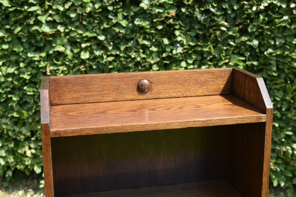 A slim, open-front Arts and Crafts oak bookcase with five shelves, cut-out side panels, and a low galleried top featuring a subtle central carved motif. View VI