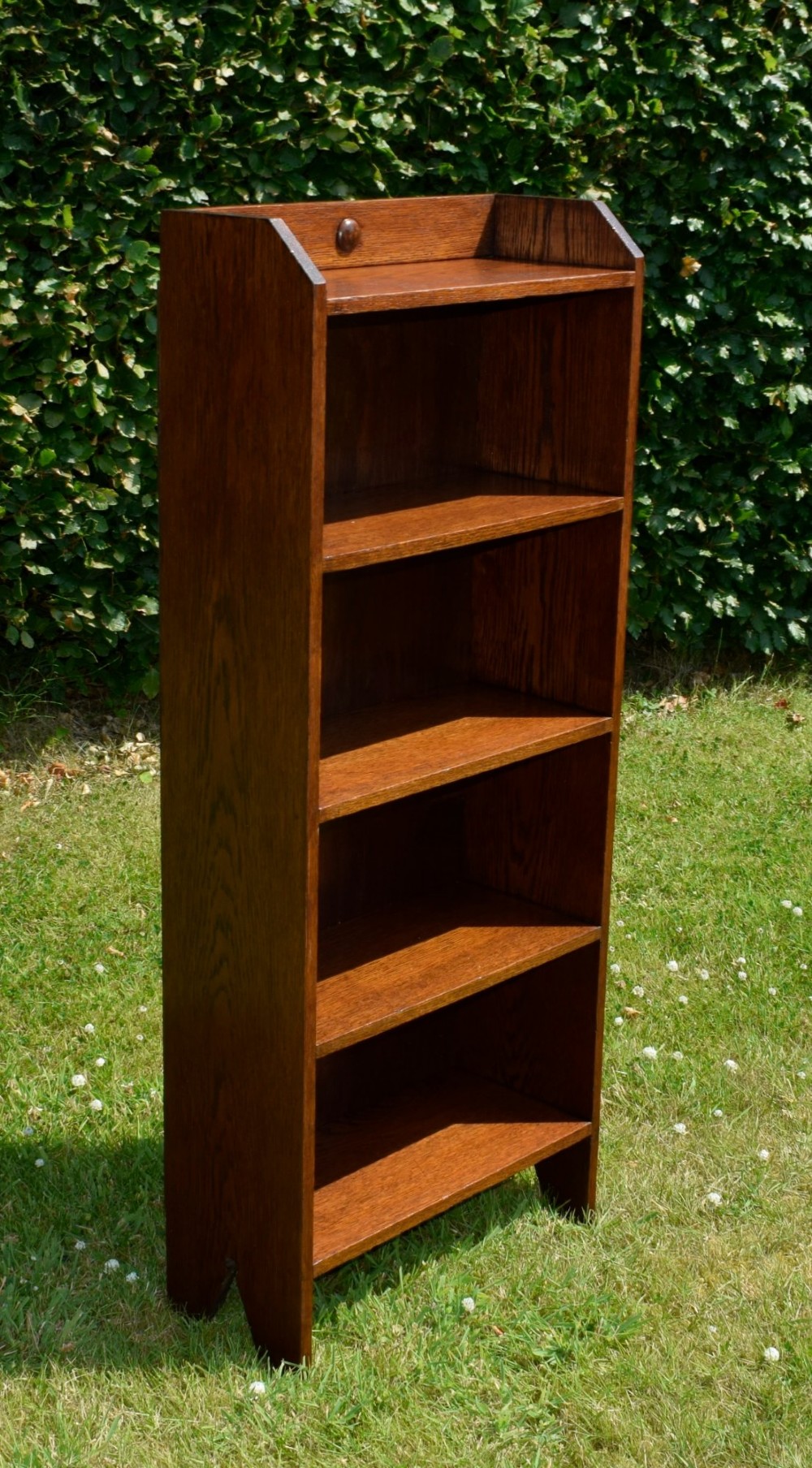 A slim, open-front Arts and Crafts oak bookcase with five shelves, cut-out side panels, and a low galleried top featuring a subtle central carved motif. View VII