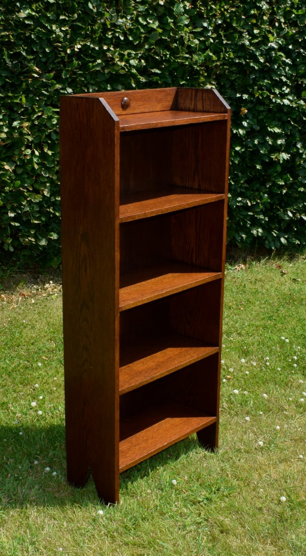 A slim, open-front Arts and Crafts oak bookcase with five shelves, cut-out side panels, and a low galleried top featuring a subtle central carved motif. View VIII