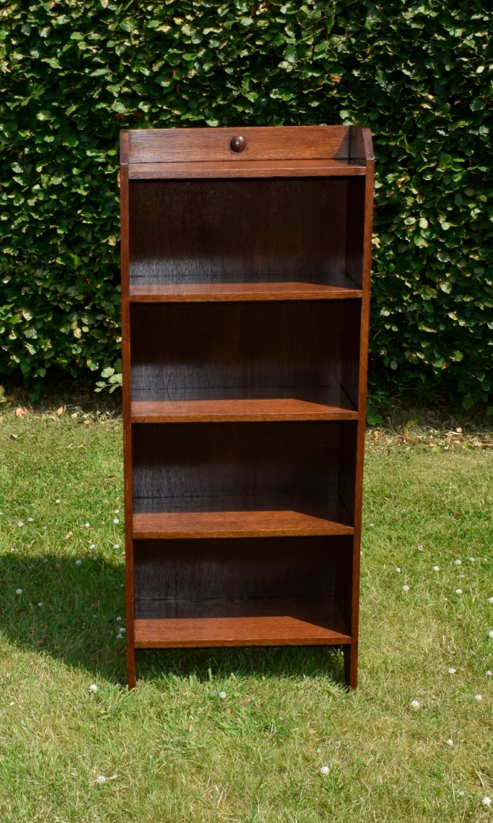 A slim, open-front Arts and Crafts oak bookcase with five shelves, cut-out side panels, and a low galleried top featuring a subtle central carved motif. View IX