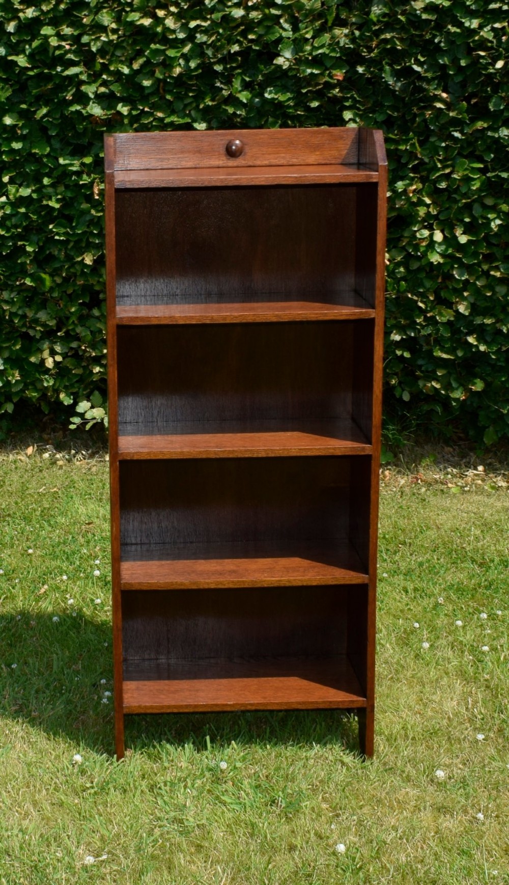 A slim, open-front Arts and Crafts oak bookcase with five shelves, cut-out side panels, and a low galleried top featuring a subtle central carved motif. View X
