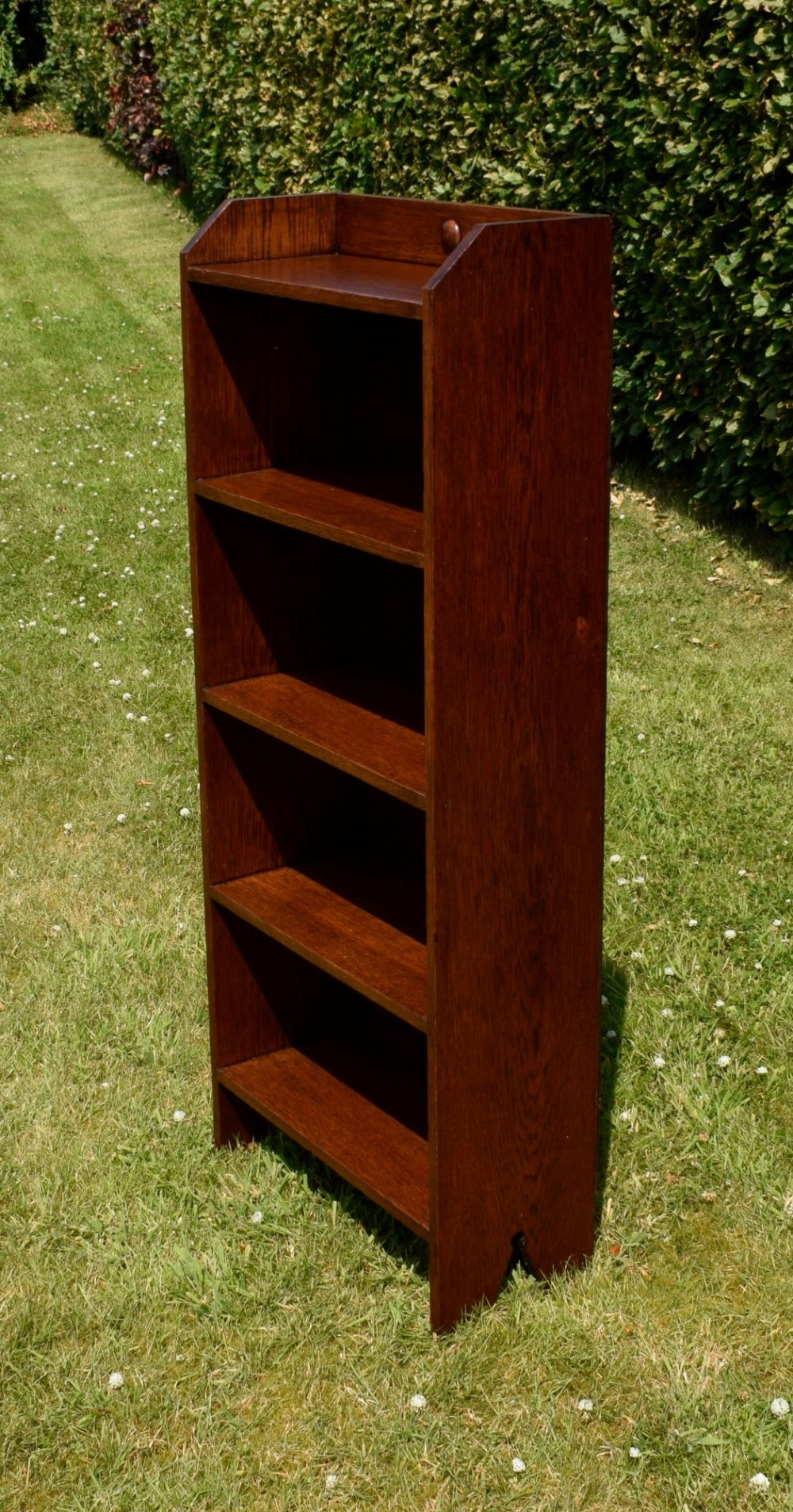A slim, open-front Arts and Crafts oak bookcase with five shelves, cut-out side panels, and a low galleried top featuring a subtle central carved motif. View XI