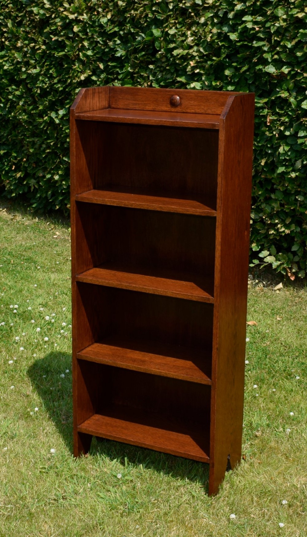 A slim, open-front Arts and Crafts oak bookcase with five shelves, cut-out side panels, and a low galleried top featuring a subtle central carved motif. View XII