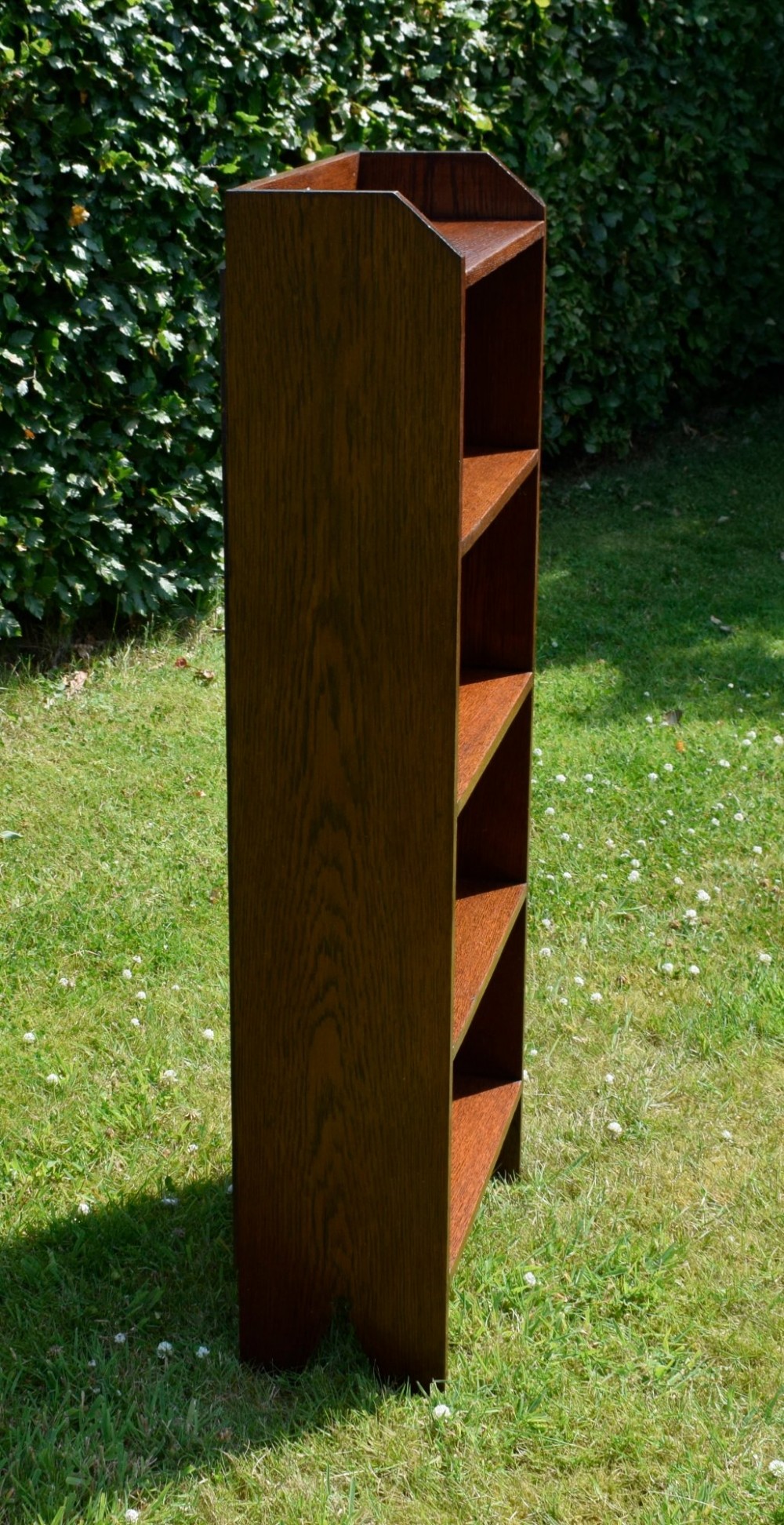 A slim, open-front Arts and Crafts oak bookcase with five shelves, cut-out side panels, and a low galleried top featuring a subtle central carved motif. View XIII