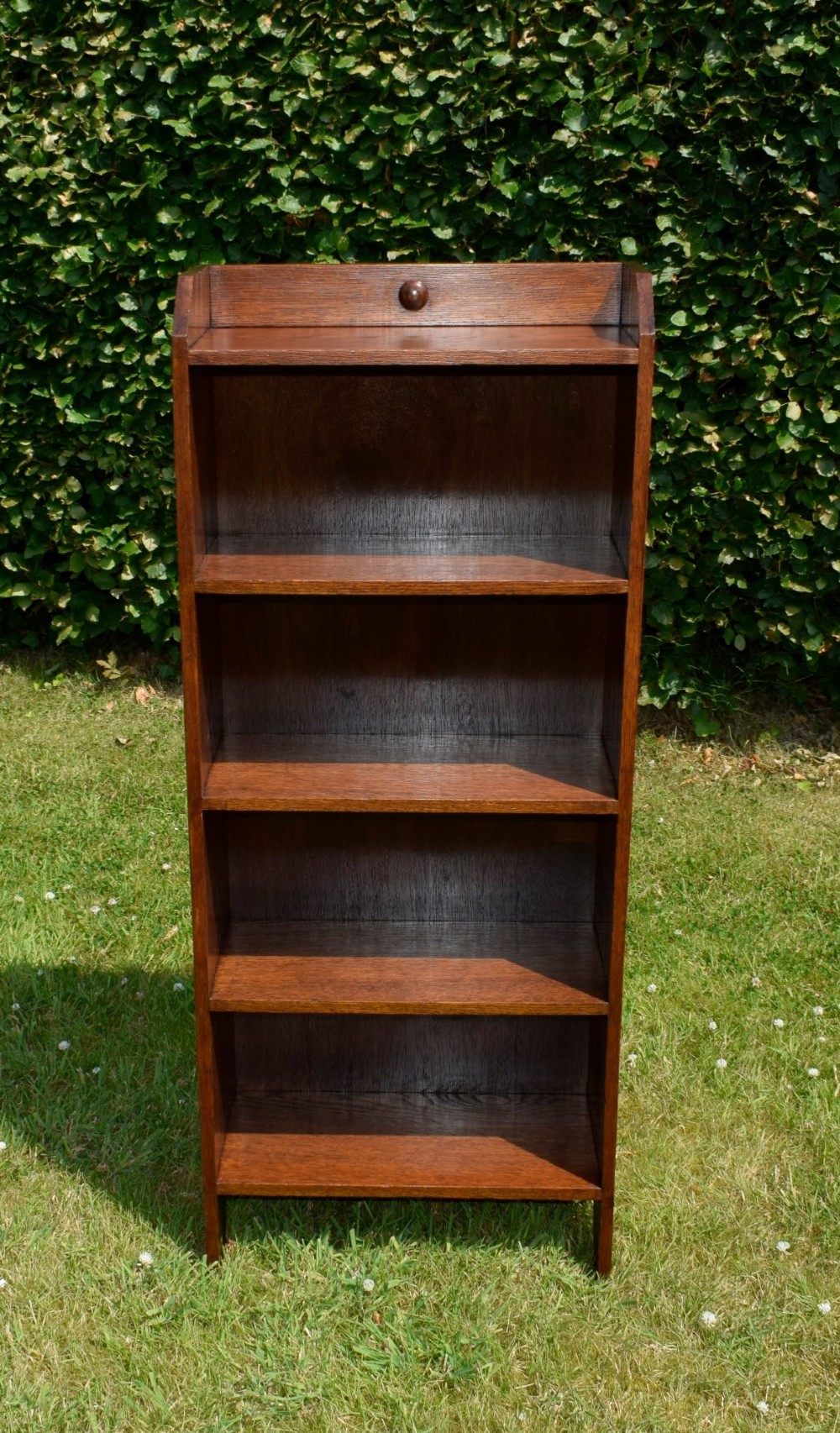 A slim, open-front Arts and Crafts oak bookcase with five shelves, cut-out side panels, and a low galleried top featuring a subtle central carved motif. View XIV
