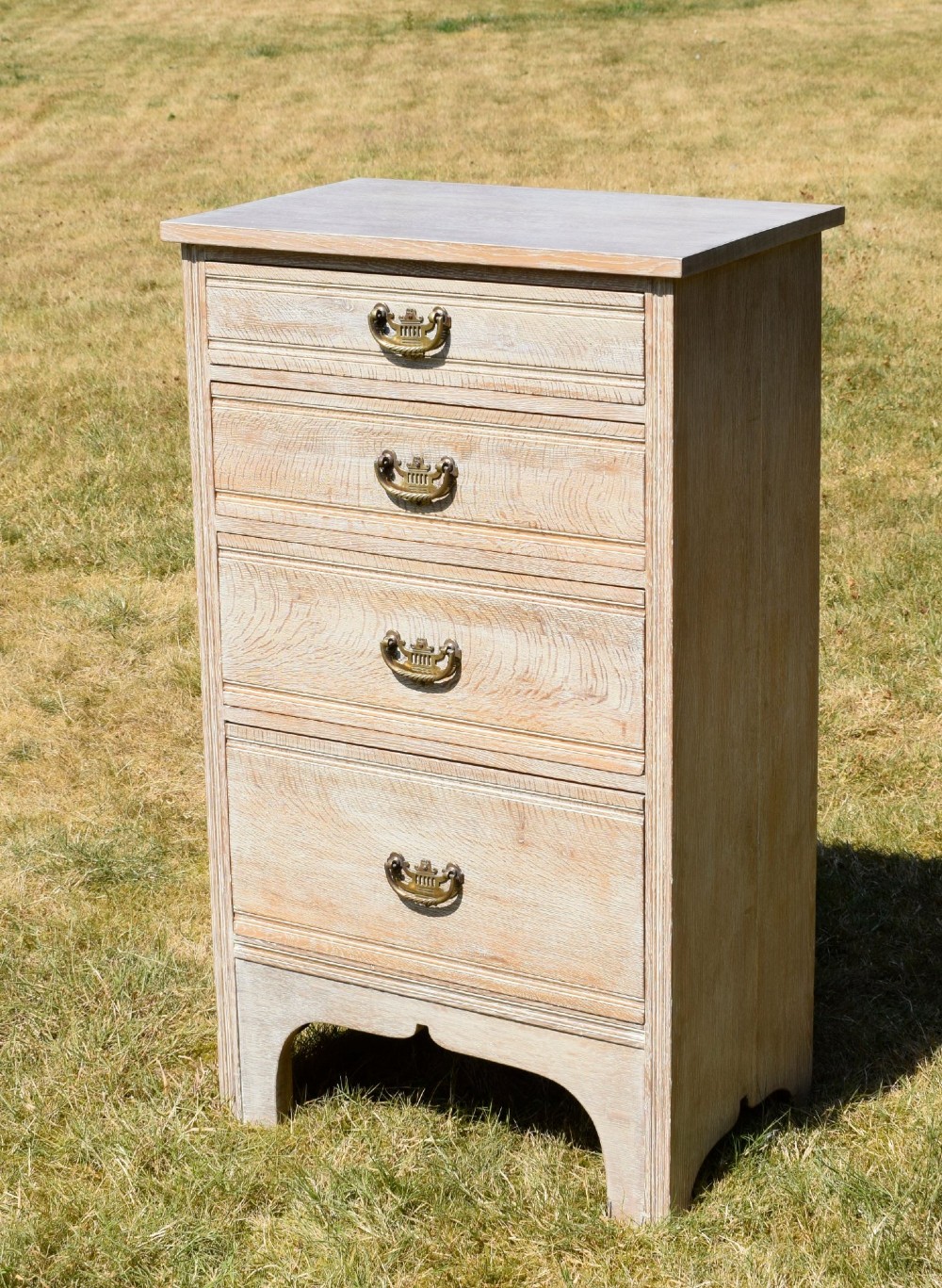 A tall Edwardian limed oak chest of drawers with four graduated drawers, brass handles, and a shaped apron base.