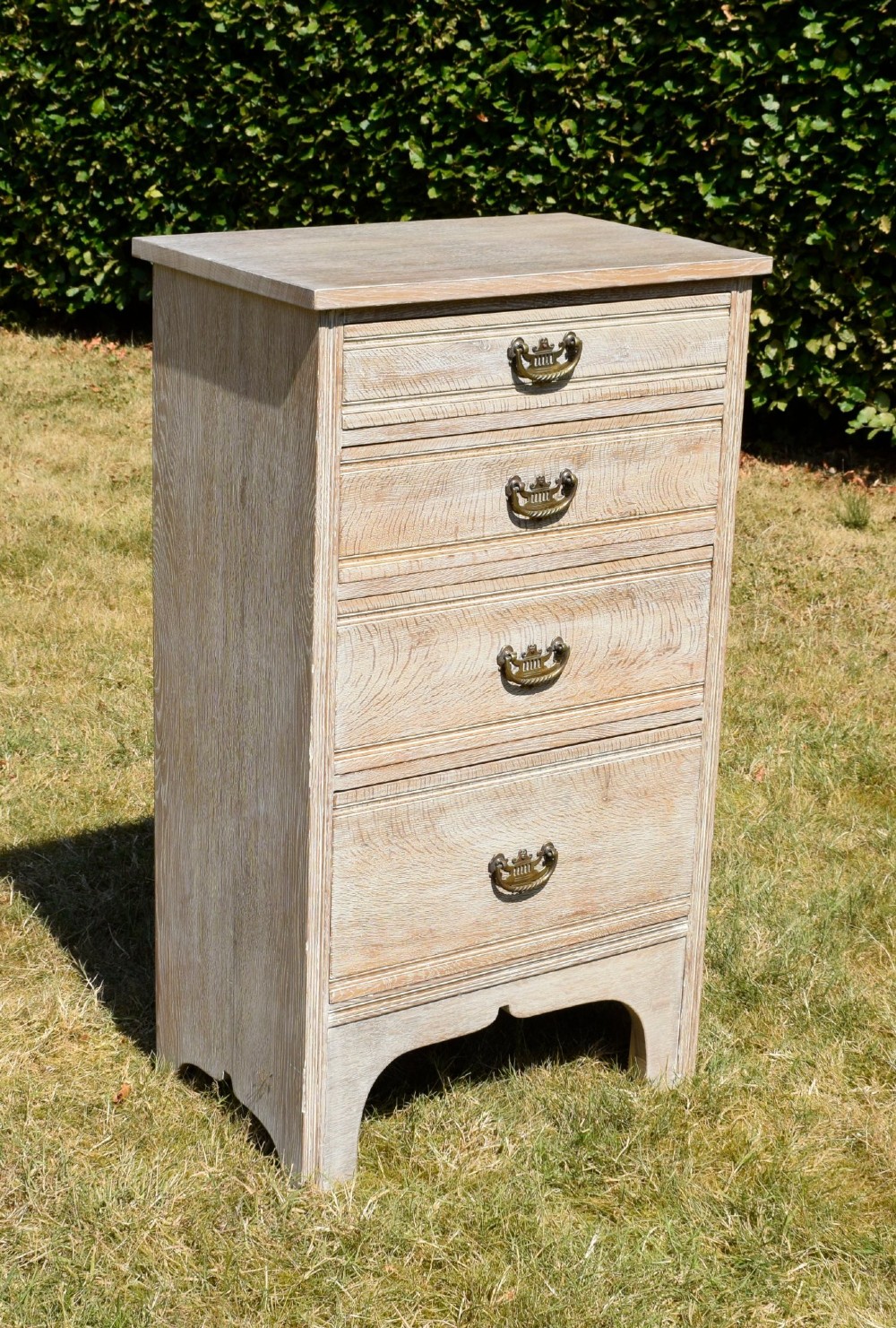 A tall Edwardian limed oak chest of drawers with four graduated drawers, brass handles, and a shaped apron base. View II