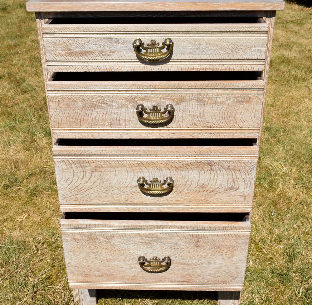 A tall Edwardian limed oak chest of drawers with four graduated drawers, brass handles, and a shaped apron base. View VI