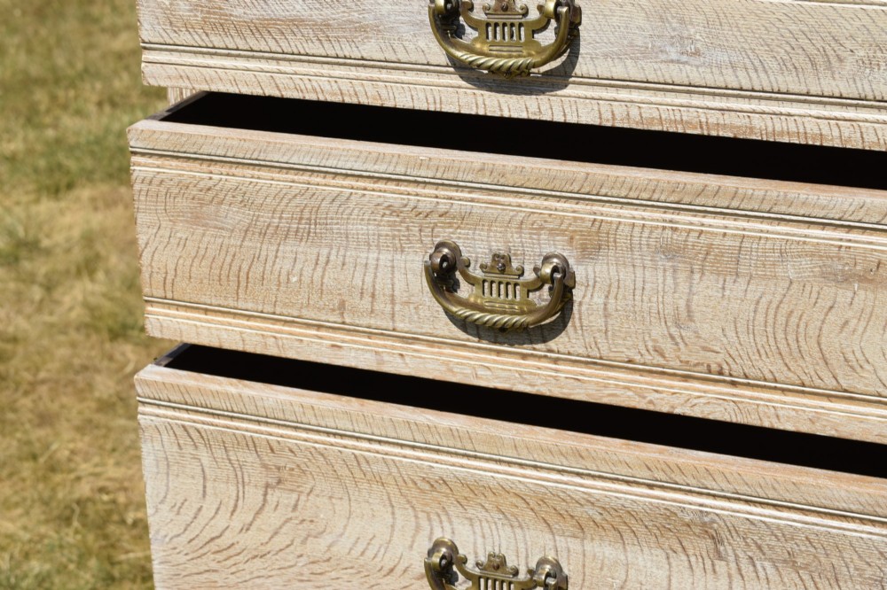 A tall Edwardian limed oak chest of drawers with four graduated drawers, brass handles, and a shaped apron base. View VII