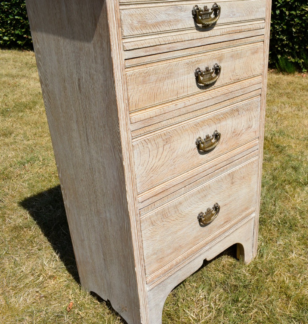 A tall Edwardian limed oak chest of drawers with four graduated drawers, brass handles, and a shaped apron base. View VIII