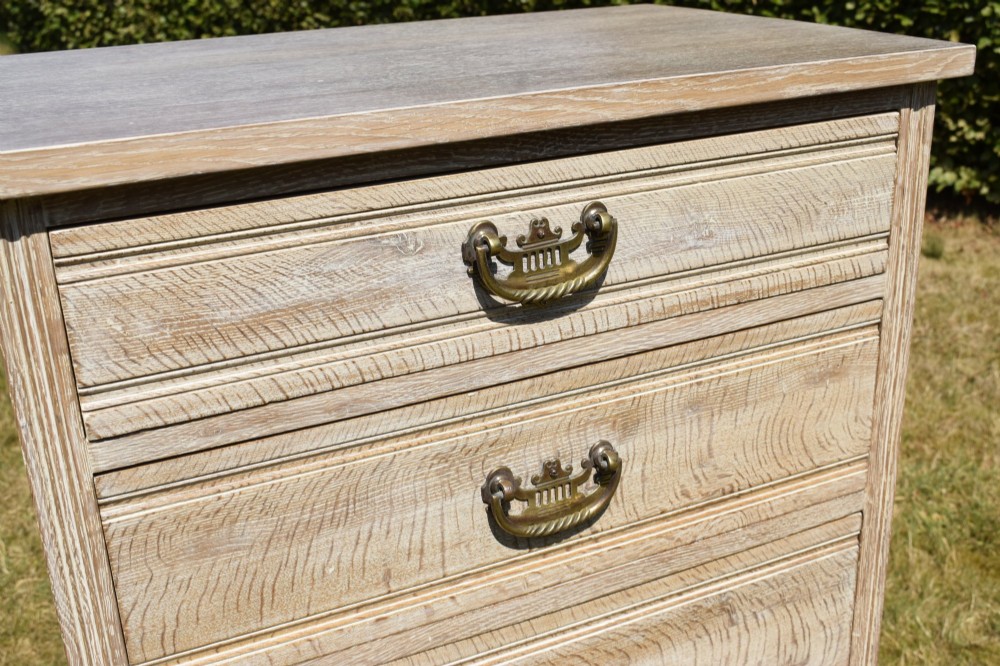 A tall Edwardian limed oak chest of drawers with four graduated drawers, brass handles, and a shaped apron base. View IX