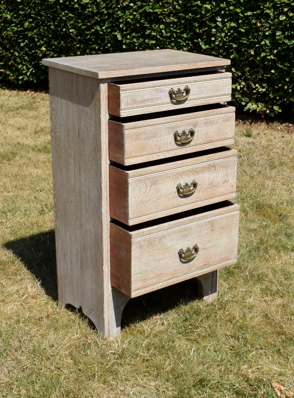 A tall Edwardian limed oak chest of drawers with four graduated drawers, brass handles, and a shaped apron base. View XI