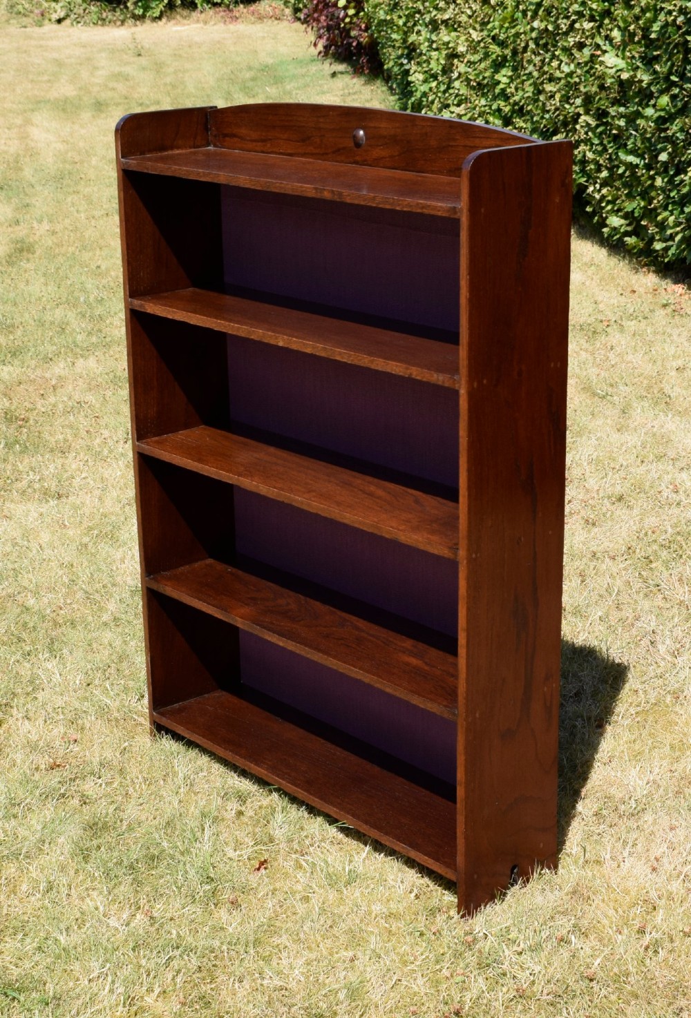 An Arts and Crafts solid oak open bookcase featuring five shelves, solid plank sides, an arched gallery top with a central decorative motif, and a purple fabric-lined back panel. View II