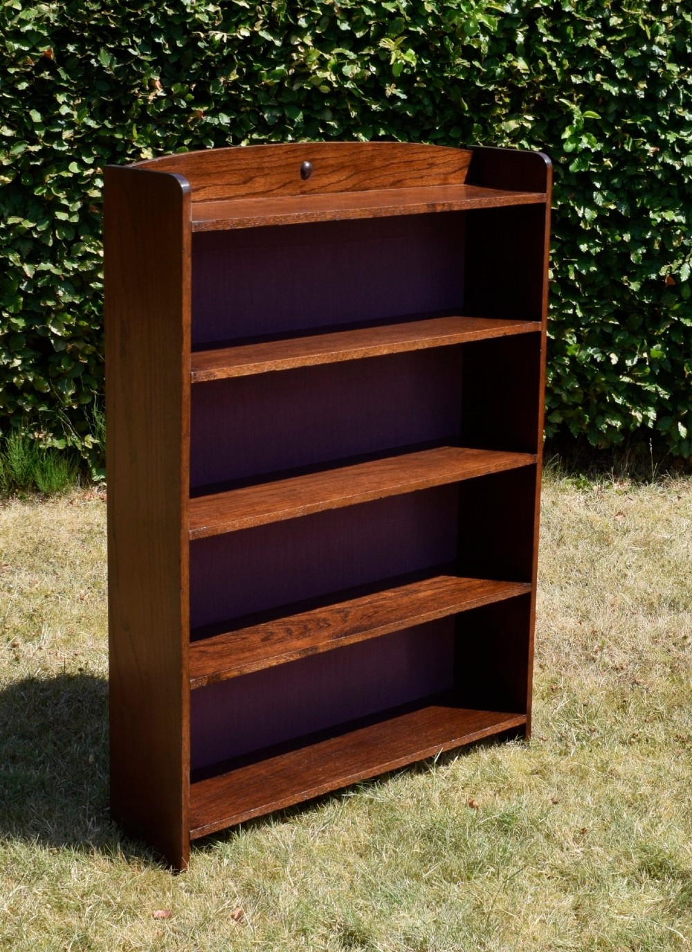 An Arts and Crafts solid oak open bookcase featuring five shelves, solid plank sides, an arched gallery top with a central decorative motif, and a purple fabric-lined back panel. View IV
