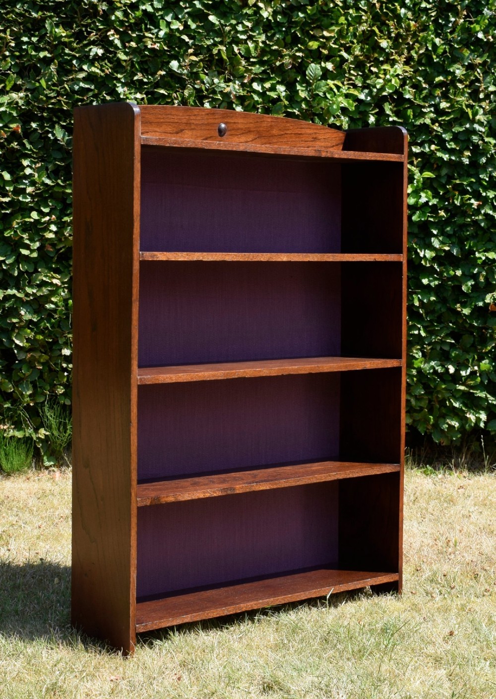 An Arts and Crafts solid oak open bookcase featuring five shelves, solid plank sides, an arched gallery top with a central decorative motif, and a purple fabric-lined back panel. View VI