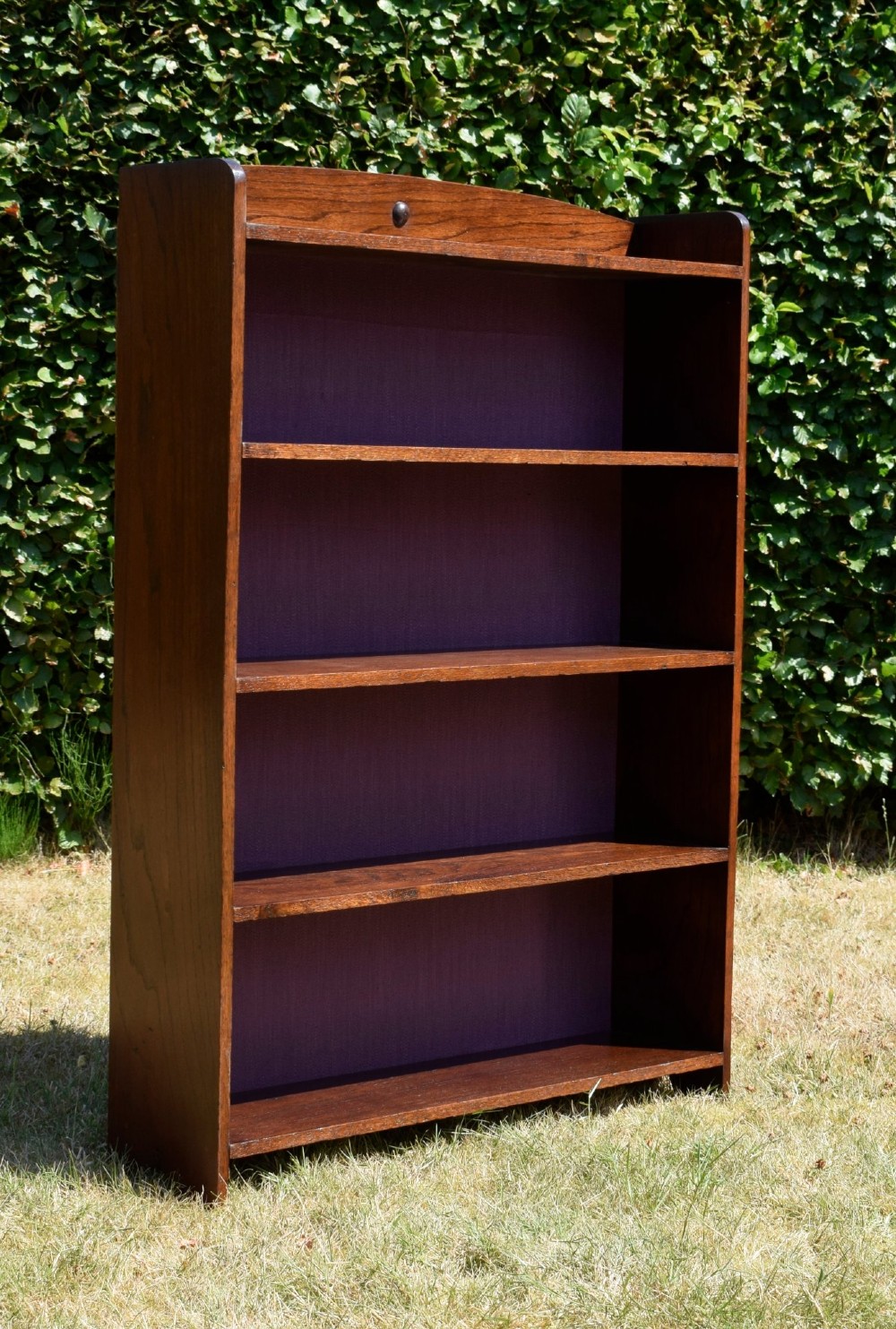 An Arts and Crafts solid oak open bookcase featuring five shelves, solid plank sides, an arched gallery top with a central decorative motif, and a purple fabric-lined back panel. View VII