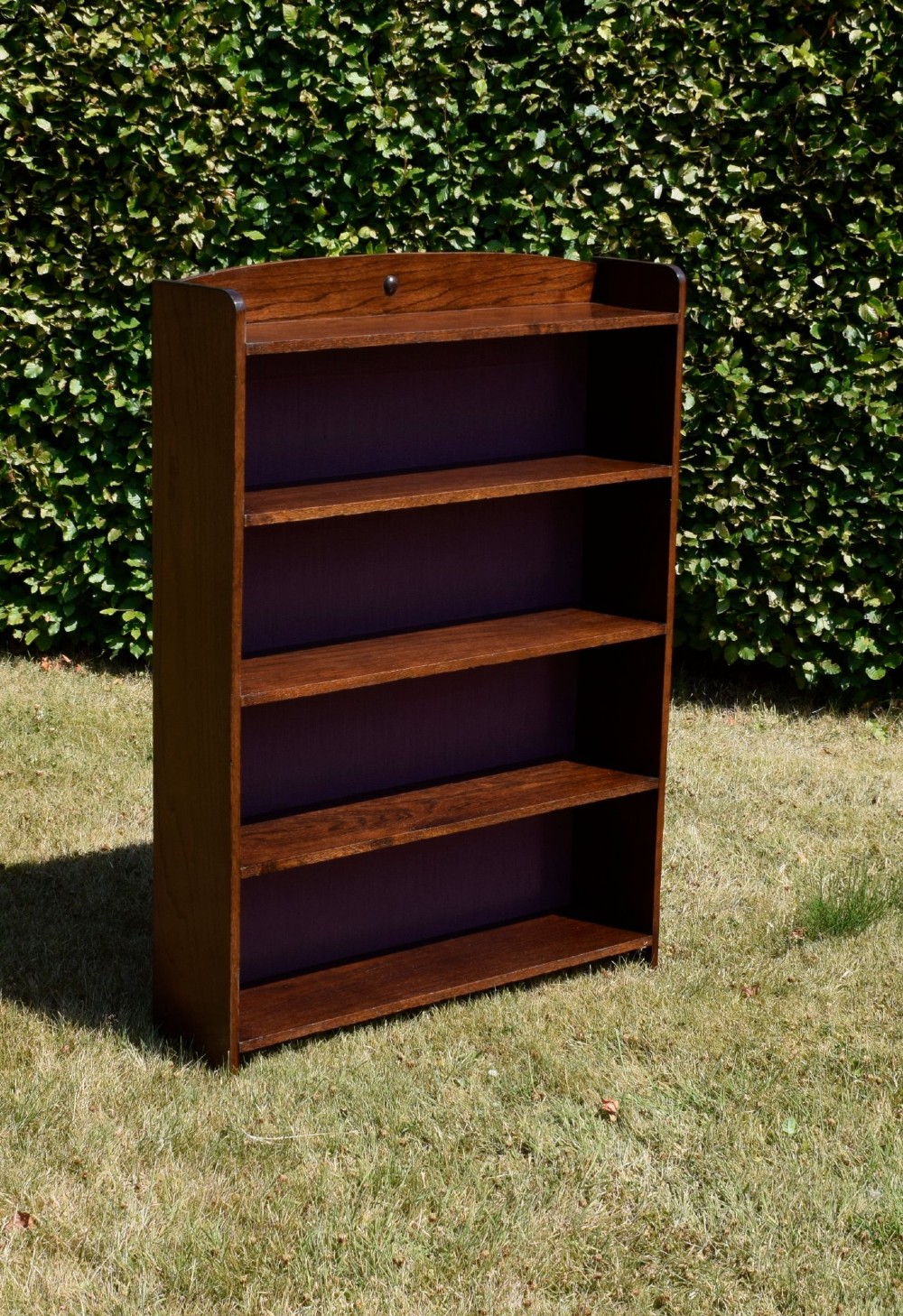 An Arts and Crafts solid oak open bookcase featuring five shelves, solid plank sides, an arched gallery top with a central decorative motif, and a purple fabric-lined back panel. View IX